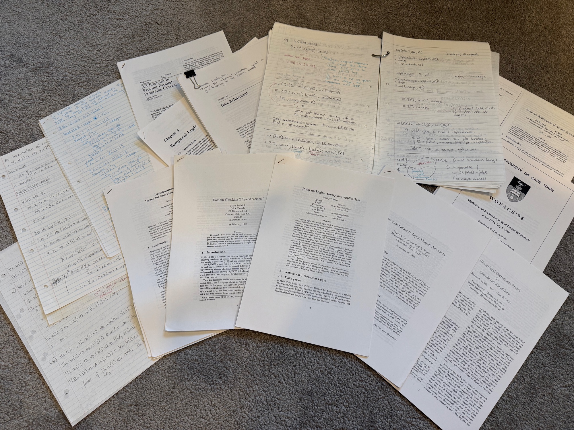 A spread of formal methods course materials on the floor: printed papers including Gries' An Exercise in Proving Parallel Programs Correct, Lynch and Tuttle's An Introduction to Input/Output Automata, chapters on temporal logic and data refinement, and pages of handwritten weakest-precondition derivations and refinement proofs.
